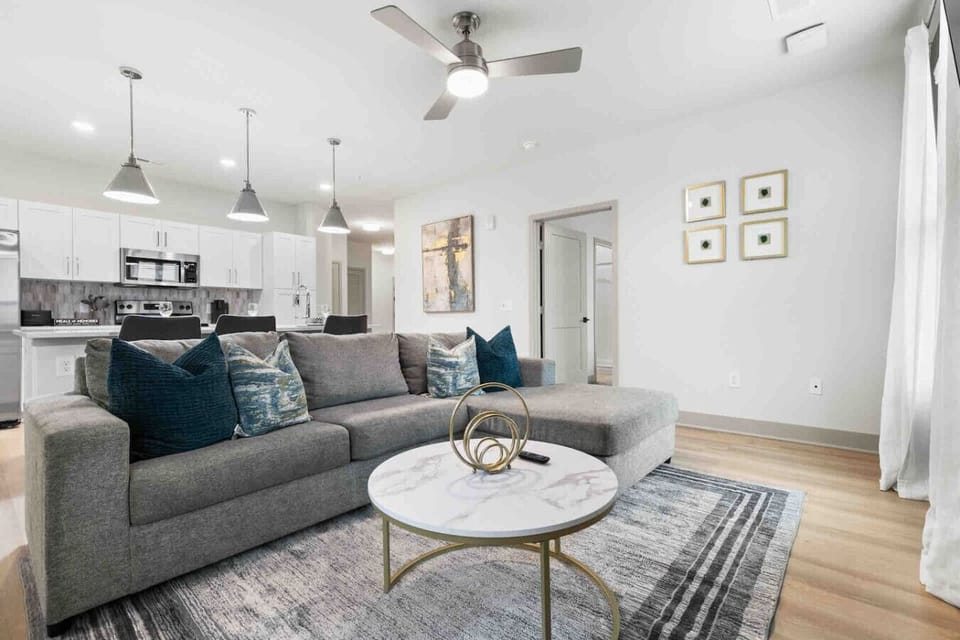 Modern Living Room with Open Kitchen- Cozy living room with plush sectional, stylish décor, and ceiling fan. Open layout flows into a bright kitchen with island seating, creating a welcoming space for relaxation, dining, and entertaining.