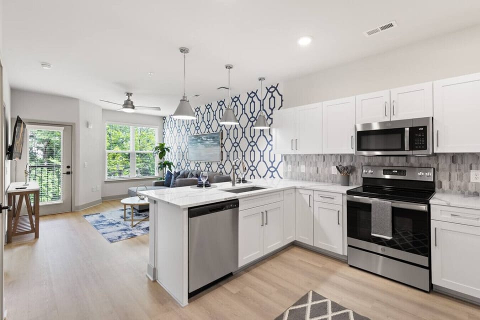 Open-Concept Kitchen & Living-Bright modern kitchen with stainless steel appliances and breakfast bar flows seamlessly into a cozy living area with natural light, chic décor, and balcony access for a stylish, functional stay.