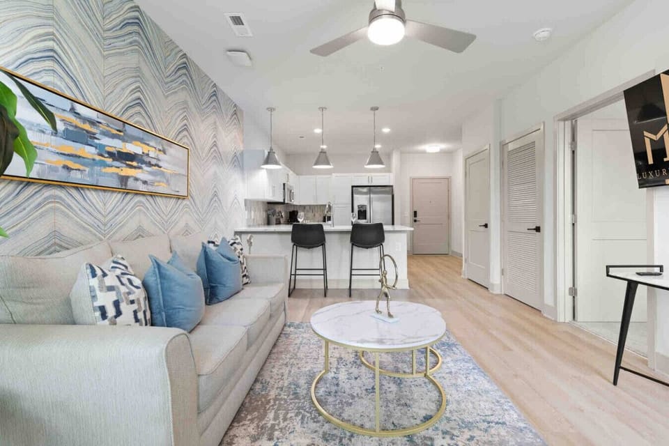 Modern Living & Kitchen Space – Open-concept design with stylish sofa, chic accent pillows, and marble coffee table. The kitchen features sleek white cabinetry, stainless steel appliances, and a breakfast bar perfect for dining or entertaining.