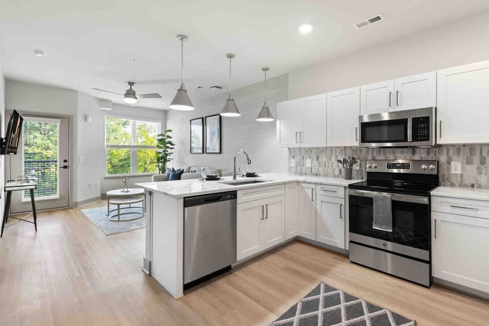 Open-Concept Kitchen & Living-Bright modern kitchen with stainless steel appliances and breakfast bar flows seamlessly into a cozy living area with natural light, chic décor, and balcony access for a stylish, functional stay.