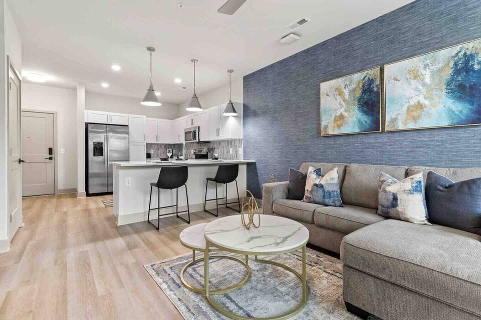 Modern Living Room – Bright and stylish space with a gray sectional sofa, chic blue accent wall, and abstract artwork. The open-concept layout flows into the kitchen with a breakfast bar, perfect for relaxation or entertaining.