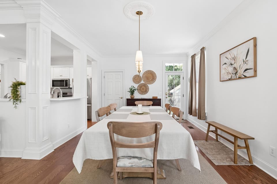 Dining room with wood floors, beige linen tablecloth, six wooden chairs, woven wall decor, and pendant light.