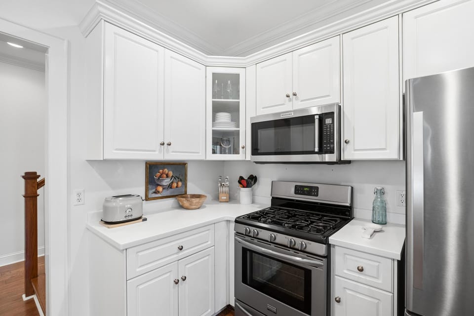 Stainless steel gas stove and microwave sit beneath white cabinetry with glass-front corner cabinet displaying dishes.
