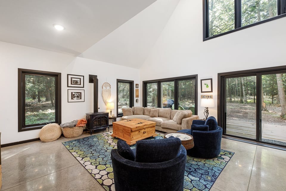 Sun-filled living room w/ soaring ceilings, cozy seating, & walls of windows framing peaceful woodland views.