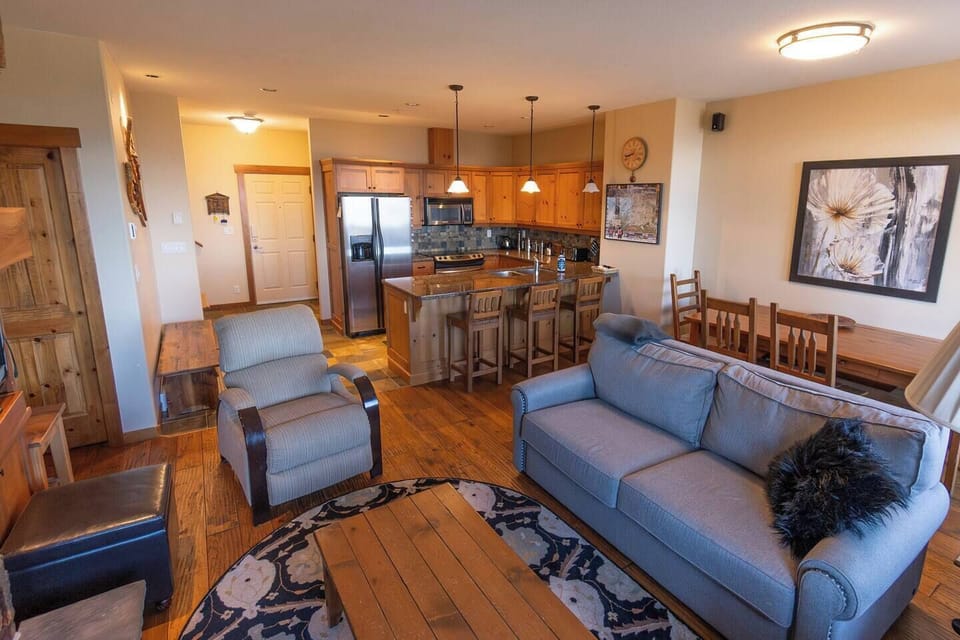 This open-concept Airbnb space features a cozy living room with a blue couch, beige armchairs, and a vibrant rug. The kitchen has stainless steel appliances, a wooden countertop, and a breakfast bar, creating a warm and inviting atmosphere.