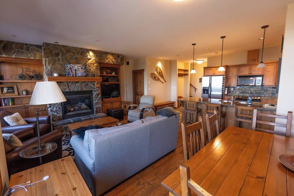 The cozy living area features a gray sofa, stone fireplace with wooden shelves, and a rustic dining table. The kitchen has stainless steel appliances, and a snowy painting adds charm to the space.
