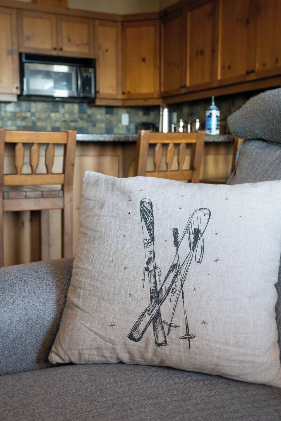 A beige pillow featuring a black-and-white print of skis and poles rests on the couch, adding a ski-themed touch to the living room. In the background, a kitchen with wooden cabinets and a bar counter is visible.