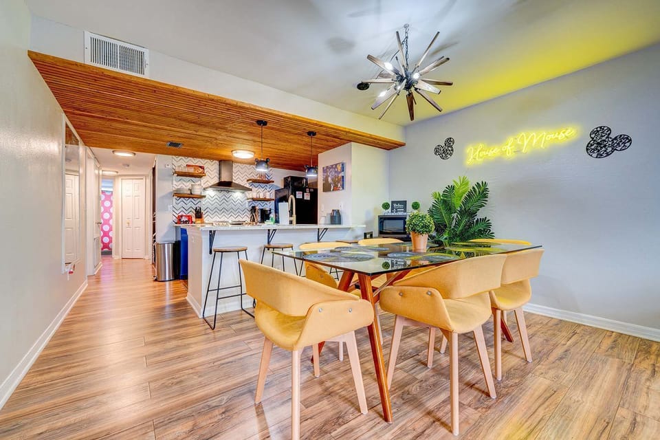 Modern dining area with a glass table and yellow chairs adjacent to an open kitchen with wood accents.