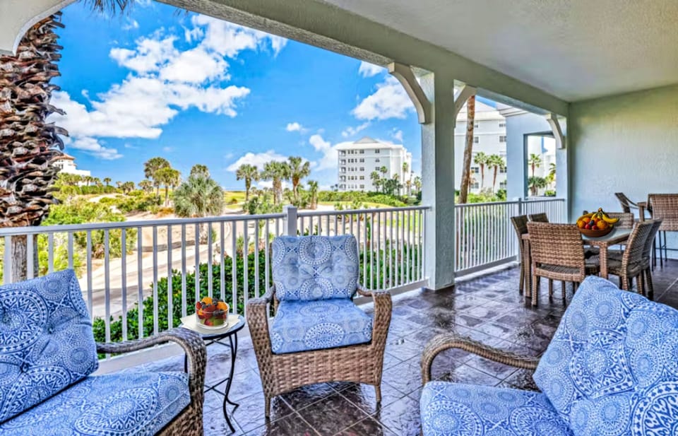 This second-floor balcony features a luxury lounge and dining area with cushioned wicker armchairs and a full outdoor table. The shaded space offers a seamless transition to the outdoors, overlooking lush palm trees and coastal architecture