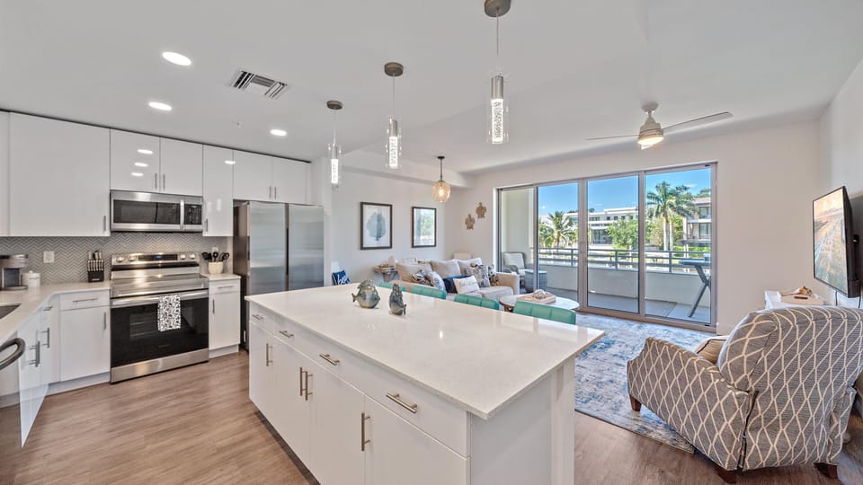 This open-concept kitchen and living space features sleek stainless-steel appliances, a spacious island, and coastal-inspired décor with views of swaying palms.