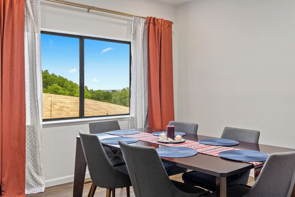 Enjoy an intimate meal at the dark wood dining table, accented by vibrant orange curtains and a bold blue tablescape, all while overlooking the serene hillside landscape.
