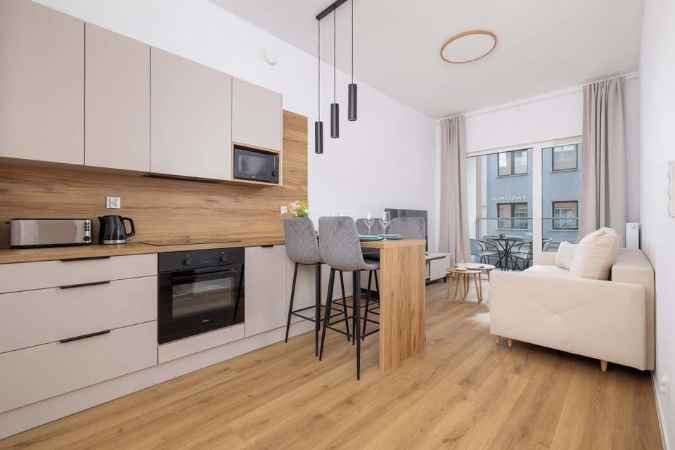 A modern kitchen with an open layout, light wood cabinetry, and a breakfast bar with stools. A dining and living area can be seen further in the background.