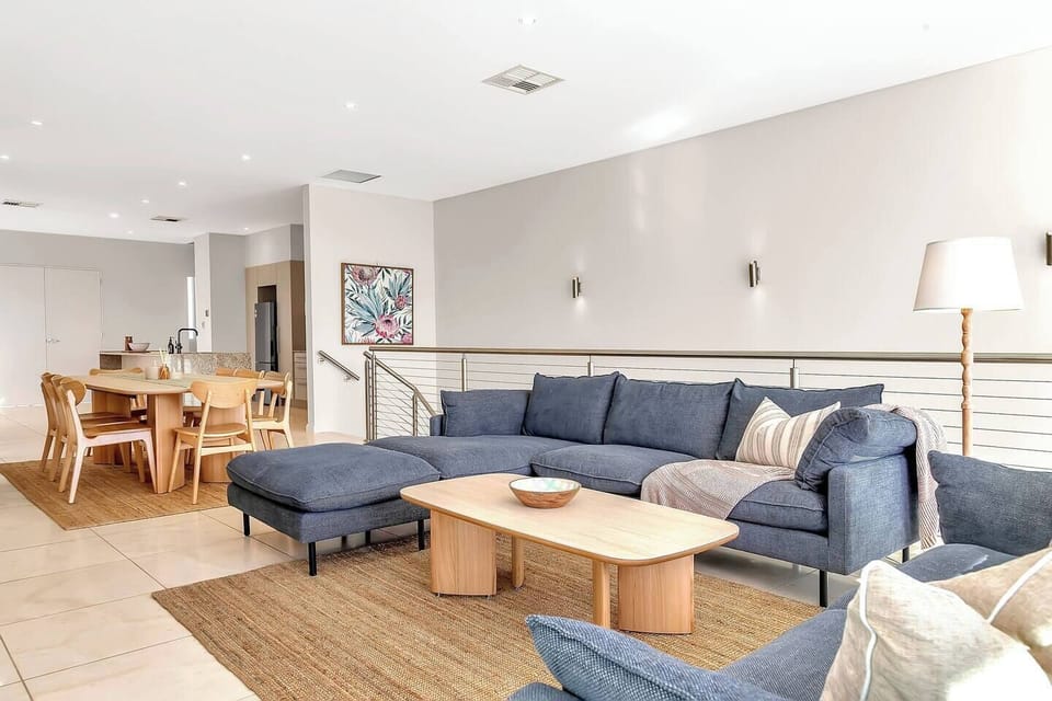 On the upper floor, the living area features plush seating and a large-screen TV, with floor-to-ceiling glass framing the coastline beyond.