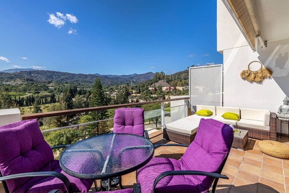 Terraza con mesa redonda de cristal, cuatro sillas acolchadas moradas, sofá modular con cojines verdes, suelo de baldosa, barandilla de cristal, acceso directo al salón y vistas amplias a colinas y vegetación en un espacio exterior funcional.
