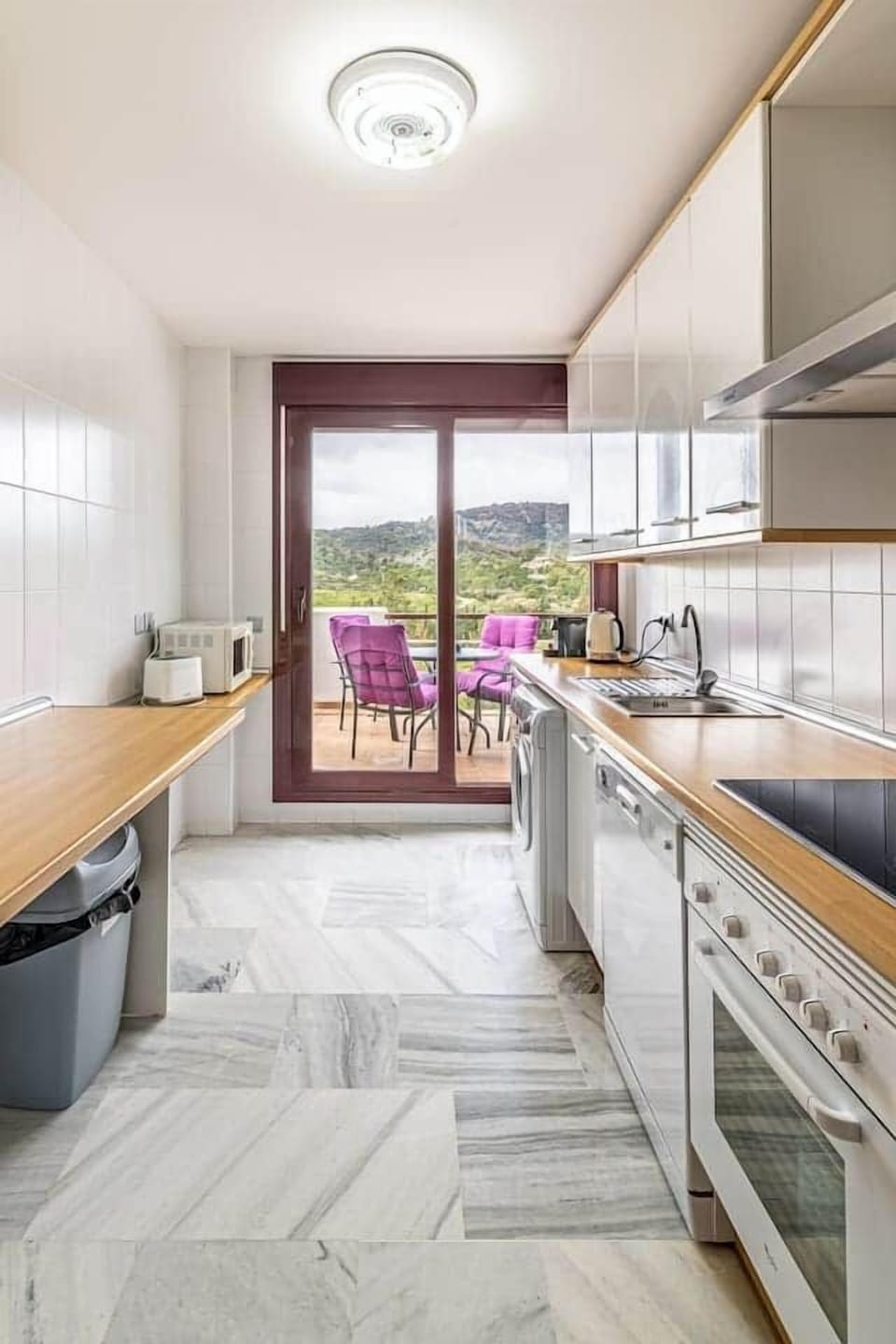 Cocina alargada con encimeras de madera, horno, vitrocerámica, campana, lavavajillas, lavadora, microondas, cafetera, nevera, armarios amplios, salida a terraza amueblada con vistas a colinas y suelo de mármol en un espacio funcional y luminoso.
