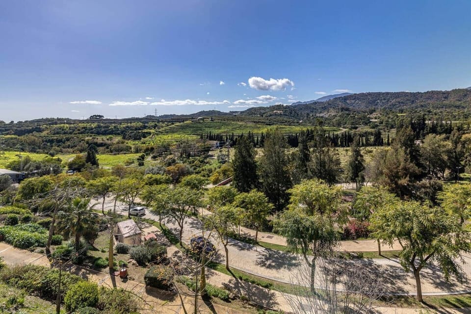 Urbanización con edificios blancos de varias plantas, caminos pavimentados, pasarela de madera sobre canal, jardines con árboles y colinas cercanas. Incluye zonas de acceso para vehículos, áreas peatonales y vistas a montañas y vegetación.
