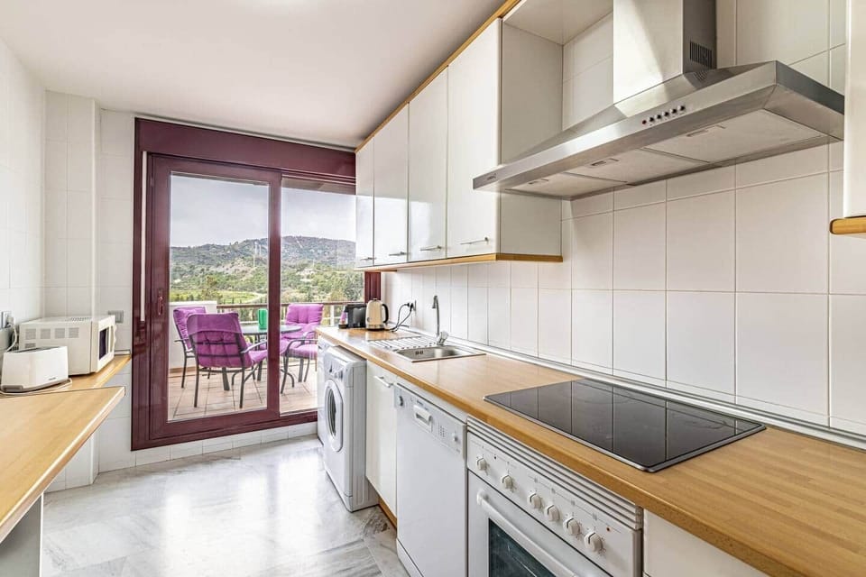 Cocina alargada con encimeras de madera, horno, vitrocerámica, campana, lavavajillas, lavadora, microondas, cafetera, nevera, armarios amplios, salida a terraza amueblada con vistas a colinas y suelo de mármol en un espacio funcional y luminoso.
