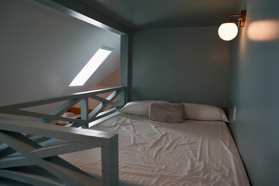 Cozy adult sized, full size custom top bunk bed tucked beneath a skylight, one of four unique bunk beds to choose from, with soft blue tones and a warm brass sconce for reading at night. The crisp linens make this nook feel like a peaceful hideaway.