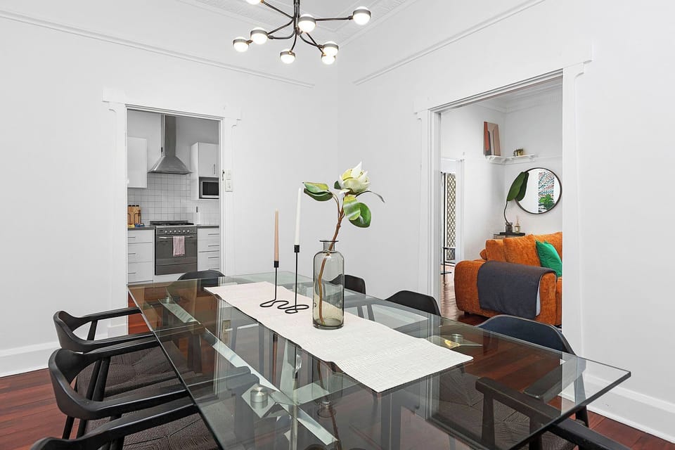 This central dining space with a sleek glass table offers seamless transitions to both the bright, modern kitchen and the vibrant living room.