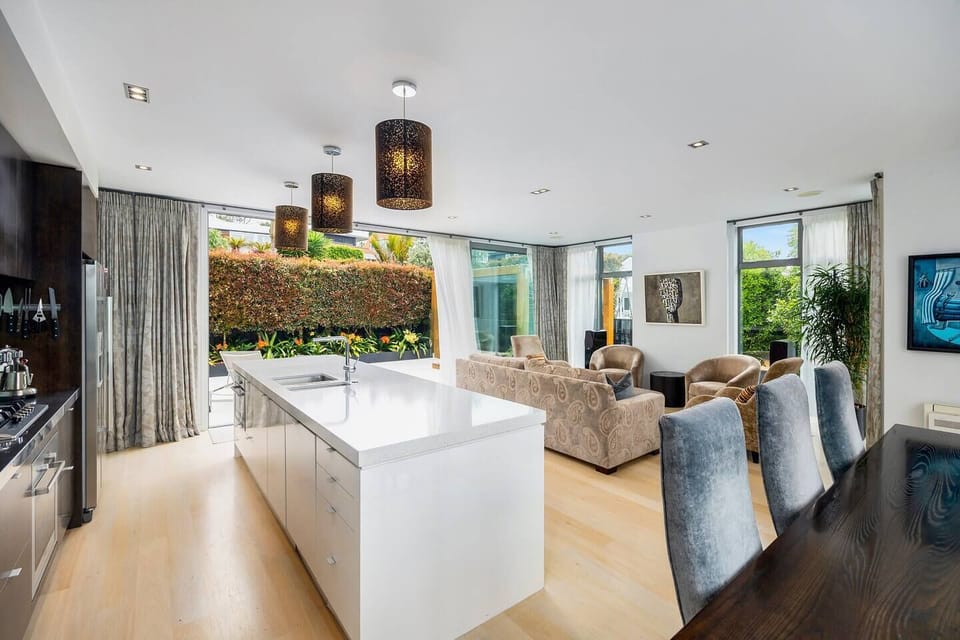 Open plan living, dining and kitchen, with doors opening out to a sunny patio with sun loungers and outdoor dining