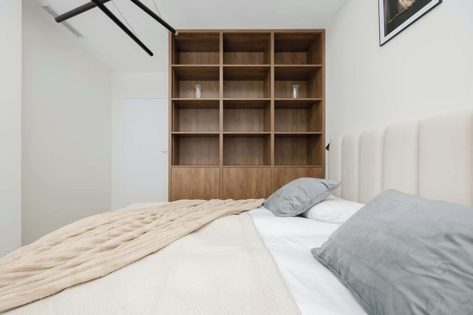 A sleek bedroom with a bed, white bedding, and natural wood flooring, with light streaming in through the windows.