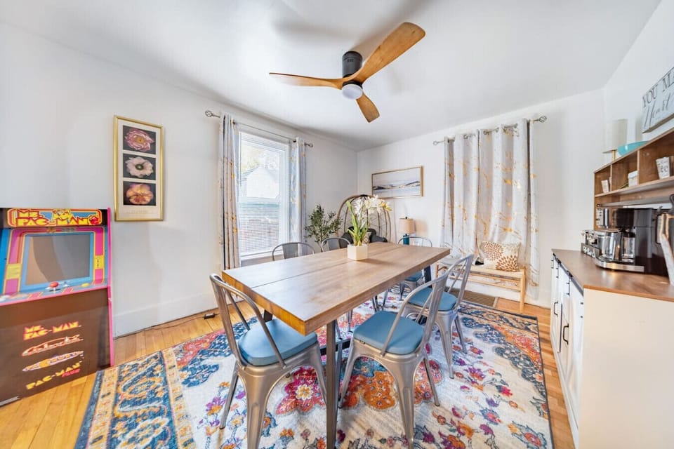 The dining area is a vibrant and inviting space featuring a large wooden harvest table paired with industrial-style metal chairs with blue cushioned seats. Set on beautiful hardwood floors, the room is anchored by a colorful, ornate area rug and a modern 