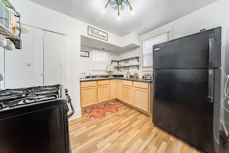 The kitchen is a bright and fully equipped space that blends classic wood cabinetry with modern black appliances and hardware. It features a double stainless steel sink, a gas range, a full-sized refrigerator, and essential countertop appliances like a to