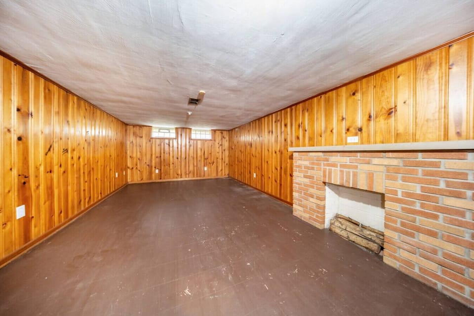 Step into this expansive bonus room, full of mid-century charm with its warm wood-paneled walls and a classic brick fireplace that serves as a cozy focal point for the space.