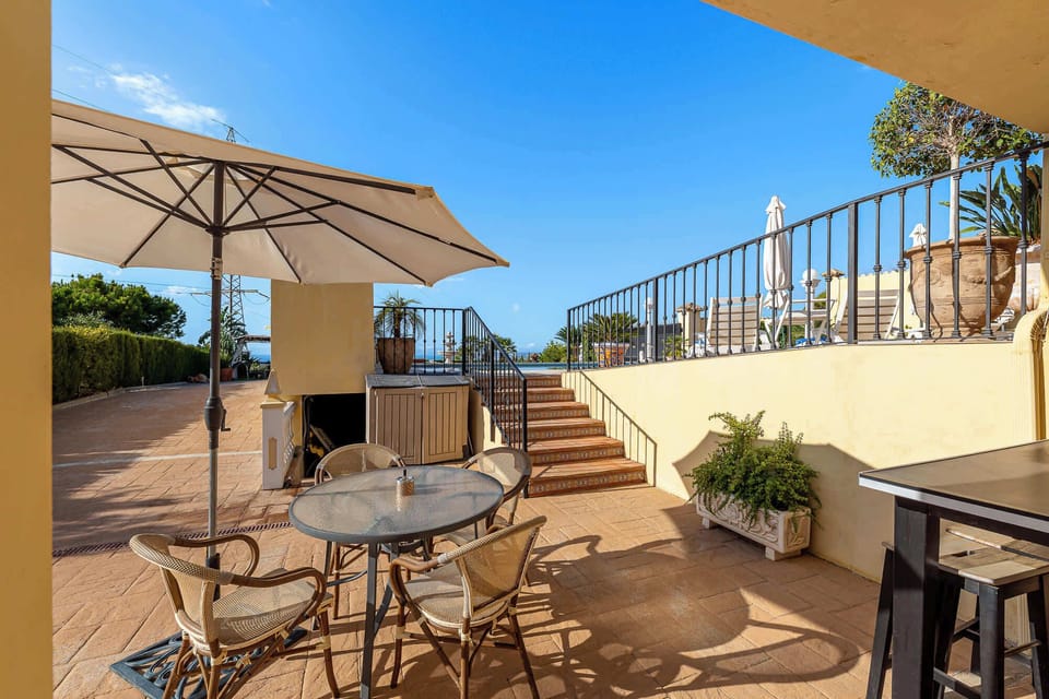 An outdoor patio with a table, chairs, and umbrella offers a relaxing space with a scenic view under a clear blue sky.