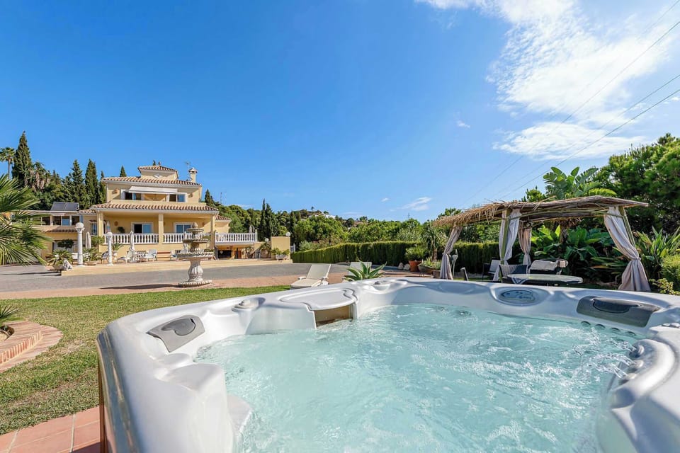 A luxurious hot tub and outdoor lounge area are set against the backdrop of a beautiful villa and clear blue sky.
