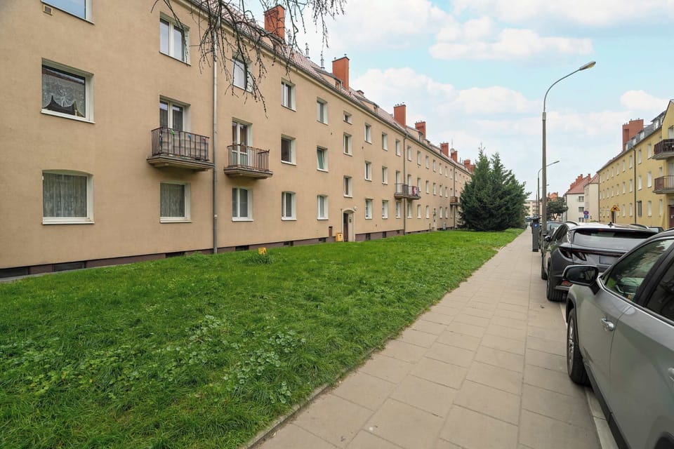 Exterior view of the apartment building along a sidewalk. The façade is beige and surrounded by a small lawn area.
