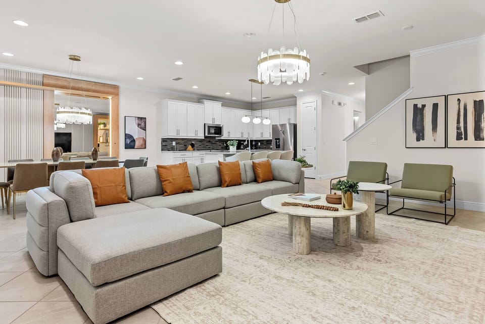 This spacious living area features a plush sectional sofa adorned with warm orange accents, inviting relaxation. Natural light floods through large windows, enhancing the modern decor and highlighting the elegant chandelier above.