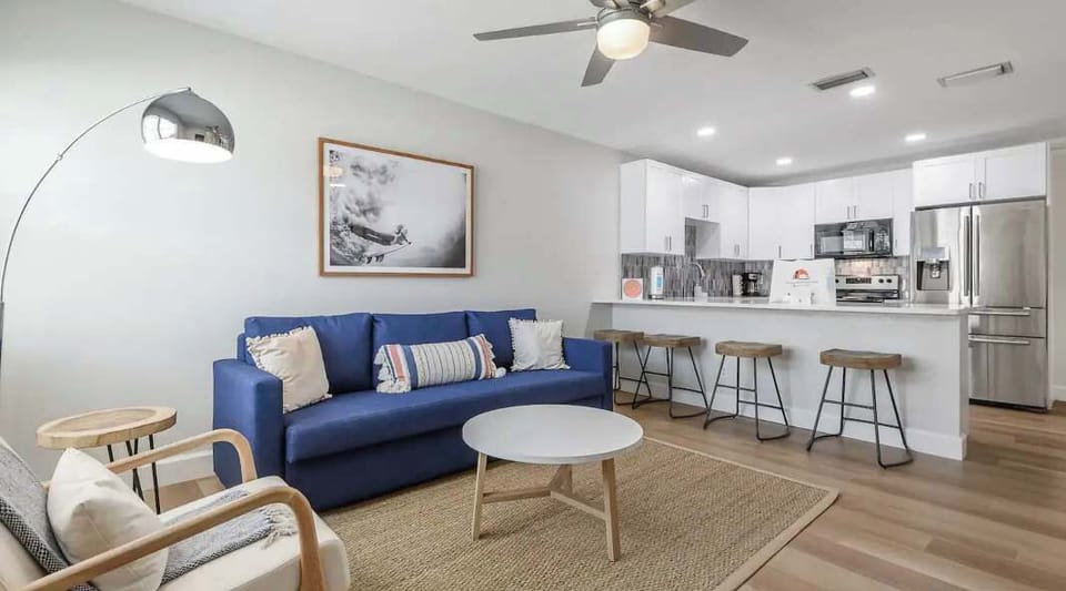 Bright and inviting open-concept living space featuring a bold blue sofa, cozy seating, and a modern kitchen with stainless steel appliances. Perfect for relaxing, dining, or entertaining in effortless coastal comfort.