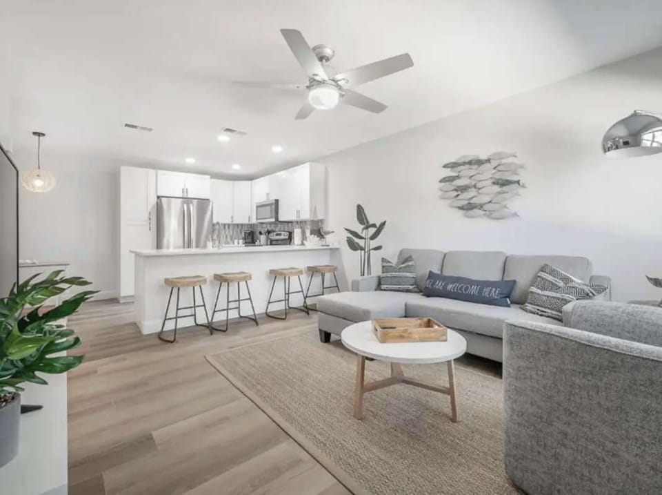 Bright and modern open-concept living space featuring a cozy sectional, sleek kitchen with bar seating, and stylish coastal décor. Natural light, warm wood floors, and soft neutrals create a welcoming retreat perfect for relaxing or entertaining.