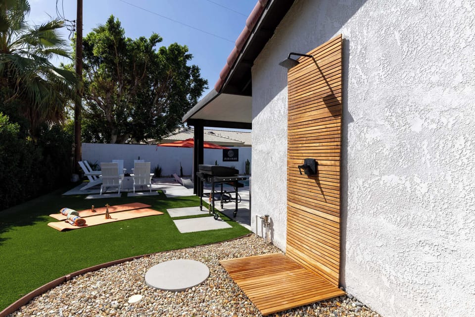 Bright outdoor teak rain shower with wood privacy screen, yoga lawn & gas grill patio beyond — rinse off and go again