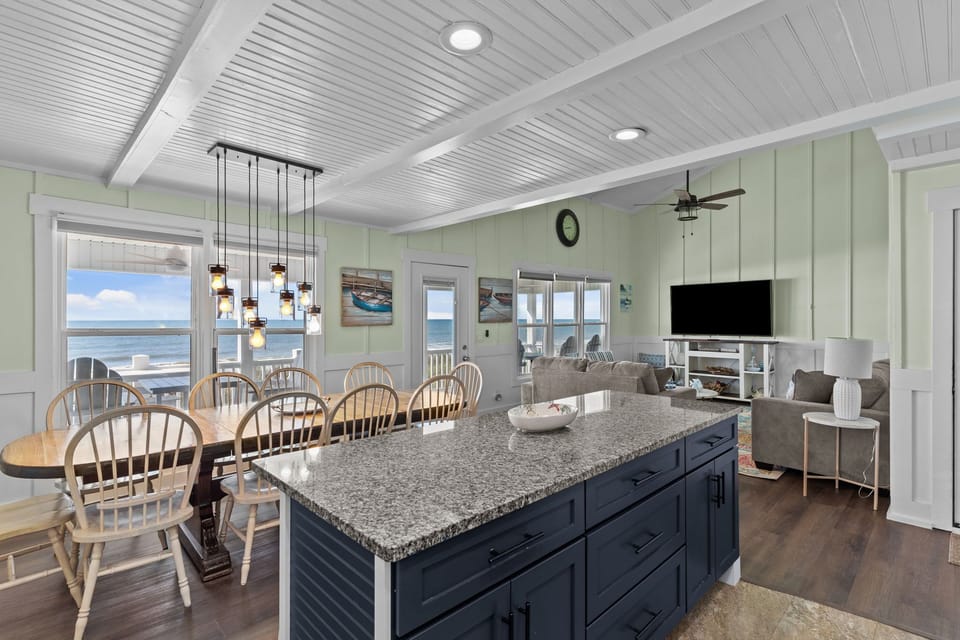 Open kitchen with island seating, granite counters, stainless appliances, tile backsplash, white cabinets, pendant lights, long dining table, chairs, wall art, and windows showing ocean and deck.