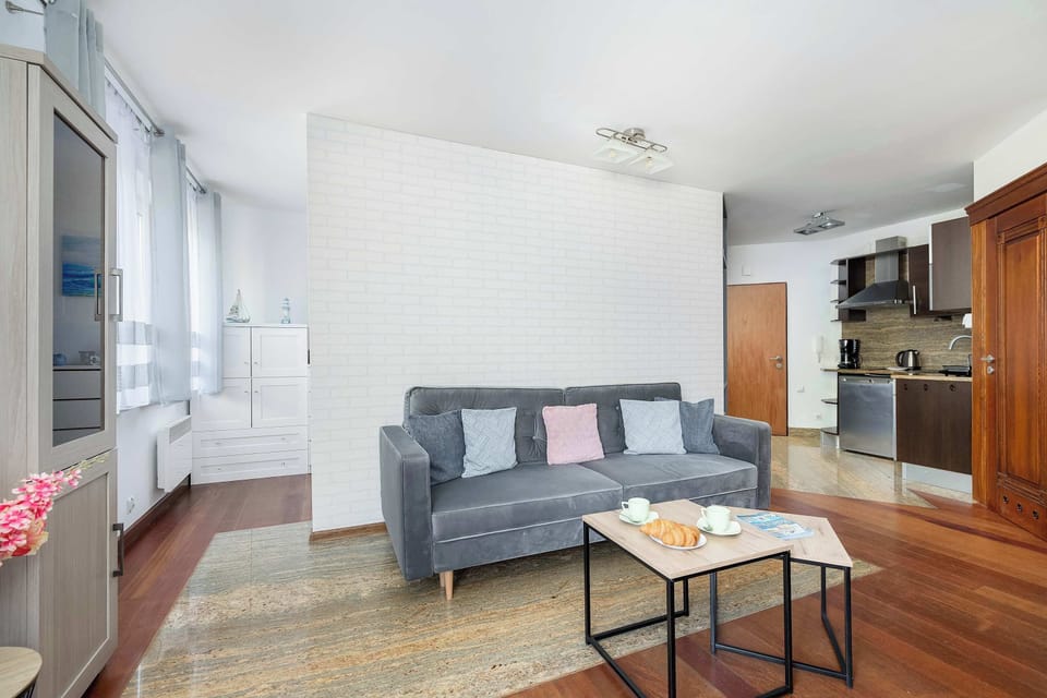 A spacious living room with modern furniture, including a gray couch and wooden floors. Large windows and light décor create an airy, welcoming feel.