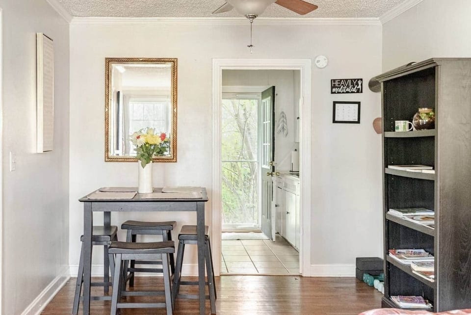 A quaint dining area with a small breakfast nook, fresh flowers, and a mirror reflecting natural light. The nearby bookshelf and hardwood floors add charm, while the open view to the back entryway creates a bright, airy feel for your stay.