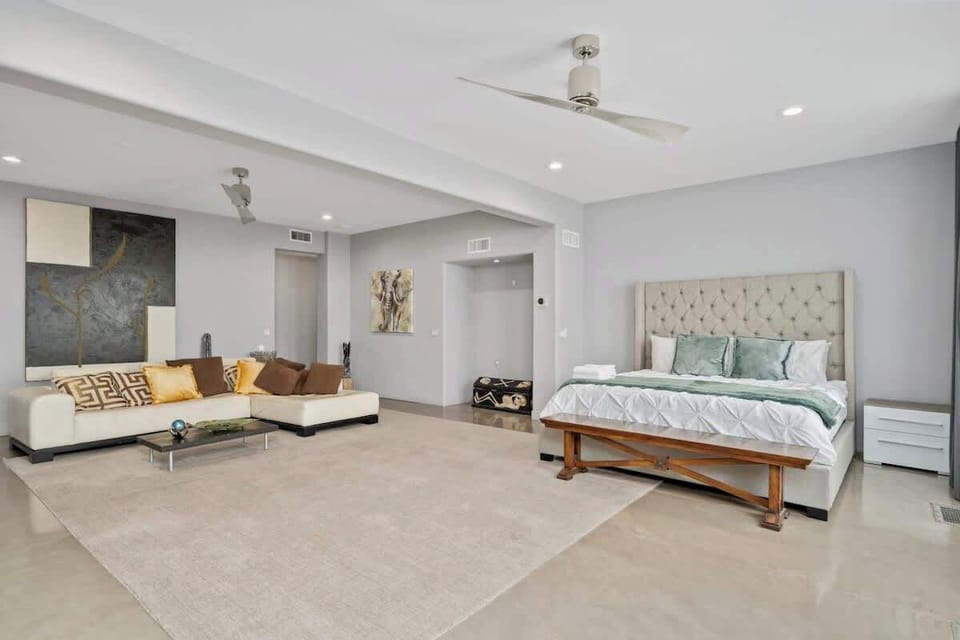This beautifully designed bedroom features a neutral-colored sectional sofa and a king-sized bed with a tufted headboard for a touch of elegance. Wide windows with sheer curtains allow for soft light, while blackout curtains cater to day sleepers.