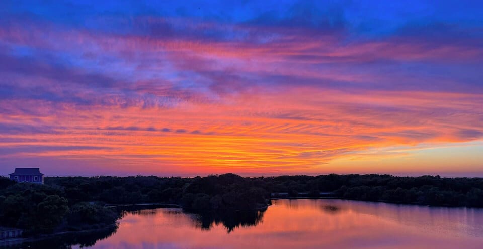 The property deck overlooks a peaceful waterway, framed by natural coastal vegetation and spectacular sunsets painting the sky in brilliant orange and purple hues.