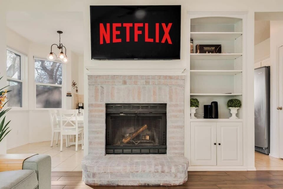 This space features a decorative white brick fireplace with a mounted 58" Smart TV, built-in shelves with greenery and storage, and a bright dining area in the background. Cozy and modern, it’s perfect for relaxing and entertaining!