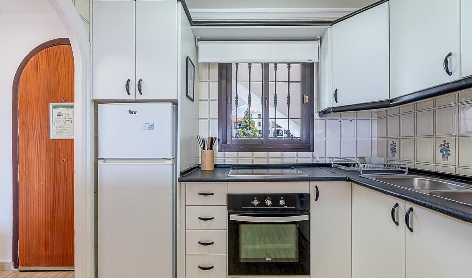 Bright, functional kitchen with white cabinetry, dark worktops, and full-size stainless steel refrigerator. Features built-in oven with ceramic hob, extractor hood, and convenient window for natural light. Everything you need for comfortable self-catering during your stay.