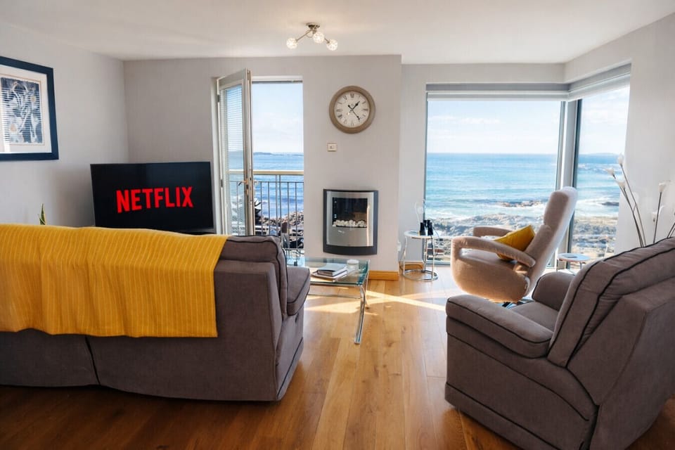 A sitting area with sofas, blanket throws, a wall clock, an electric fireplace unit, and large windows looking directly towards the coastline.