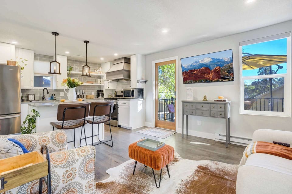 Light filled living area opens to the kitchen, with patterned chairs, a sofa, and an orange ottoman creating a comfortable place to gather while bar stools at the counter keep everyone connected during meals or conversation.