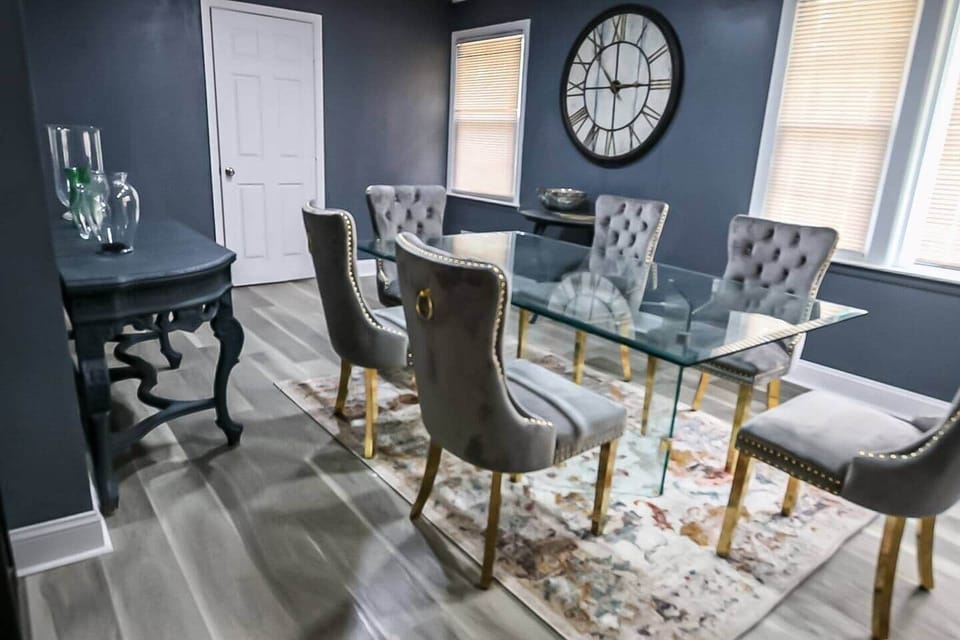 Pull up a tufted velvet chair with gleaming gold legs to a floating glass table—set atop a painterly rug. A colossal wall clock on grey navy walls anchors this edgy, elegant dining retreat.
