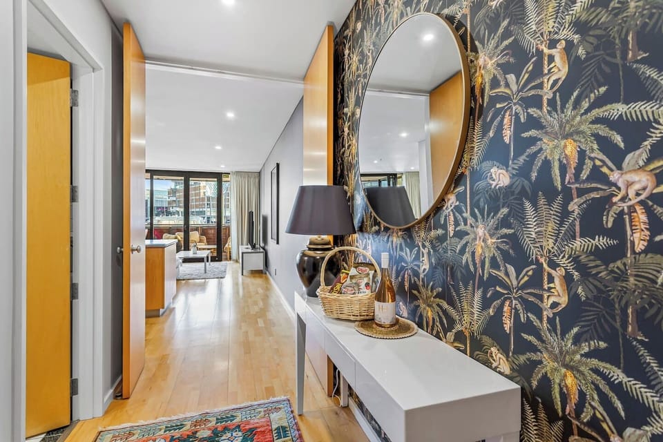The first thing you see when you walk in — a bold tropical wallpaper, welcome wine, a round mirror catching the light, and the marina glinting at the end of the hallway. A genuinely warm arrival