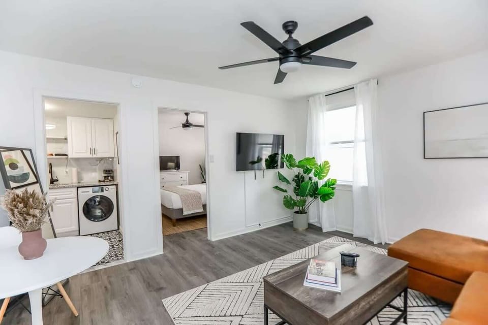 Living room with a view of the kitchen including a washer and dryer. Living room has a ceiling fan, coffee table, windows, mounted television, cafe table with chairs and a comfortable couch. 