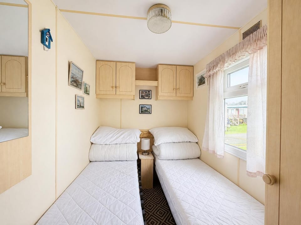 Two single beds with white quilted covers sit beneath light wood overhead cabinets and soft pink lace curtains.