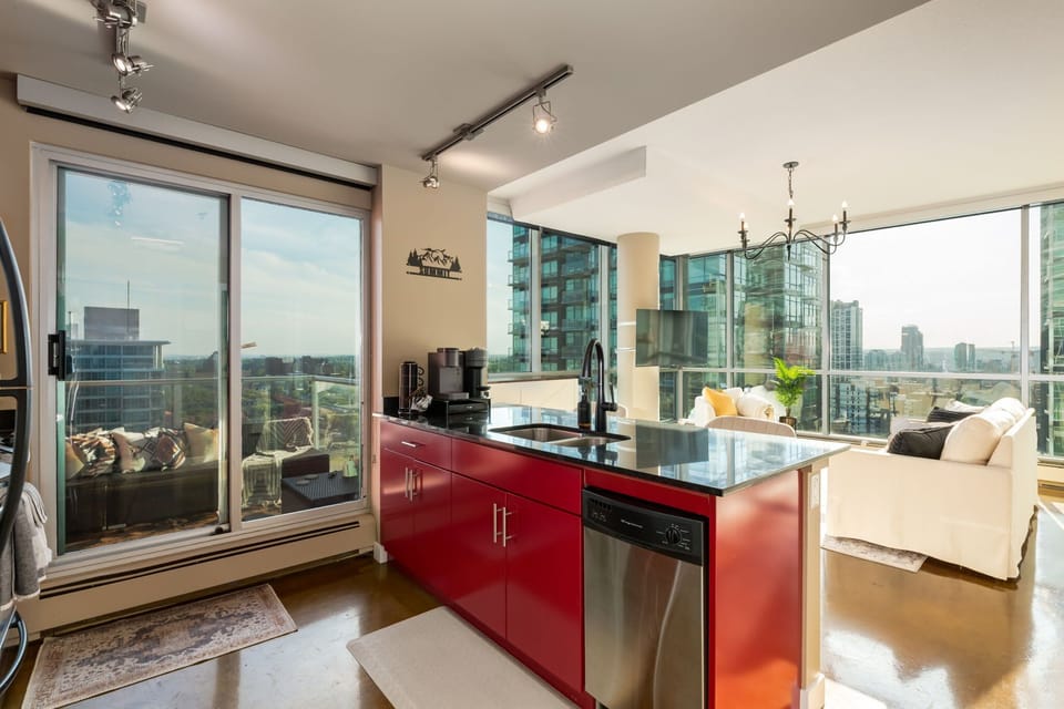 Modern condo with floor-to-ceiling windows showcasing stunning city views. Features a sleek kitchen with bold red cabinetry, granite countertops, and stainless steel appliances. The open living area flows to a private balcony.