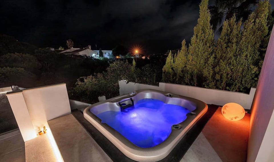 A glowing hot tub on a sleek patio, surrounded by manicured trees and candlelight, radiates luxury and calm under a cloudy night sky.